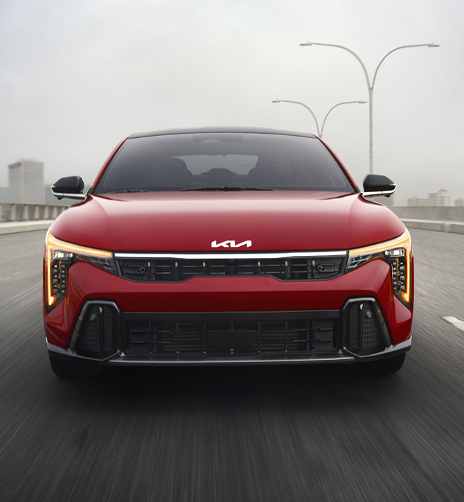 Front view of the 2026 Kia K4 sedan driving straight towards the camera, showing its windshield, hood, front grille, and amber daytime running lights with cube projector headlights on the GT-Line Turbo trim in red.